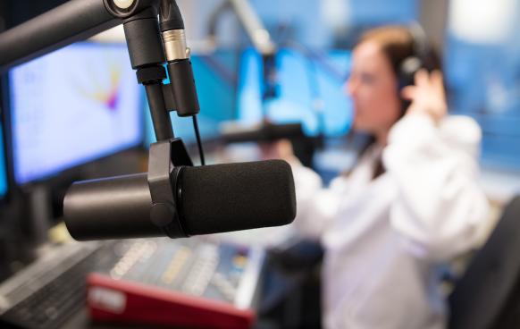 Photo of a podcasting microphone in a studio environment with a woman on headphones out of focus in the background.