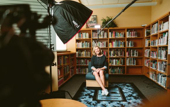 Woman sitting in a school library with a video light, camera, and boom microphone pointed at her. A setup for an interview. Photo by Sam McGhee on Unsplash