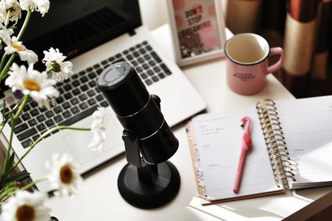 A microphone on a simple desk stand on a desk with a laptop, flowers and a notebook with a flamingo styled pen.