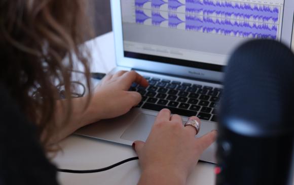 A woman at a laptop computer editing podcast audio in Audacity. A microphone is out of focus in the foreground. (Photo by CoWomen on Unsplash)